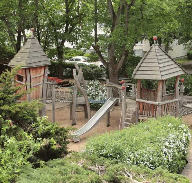 Ein hölzerner Spielplatz mit zwei Türmen, einer Rutsche und Kletterelementen inmitten von Bäumen und grünen Sträuchern.