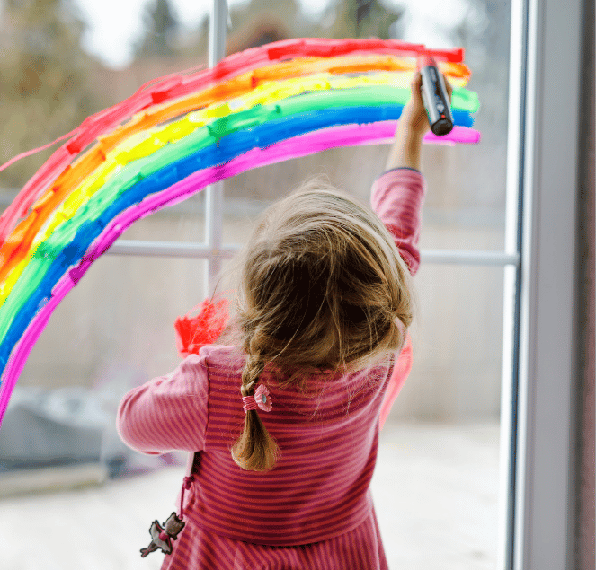 Ein kleines Kind mit Zöpfen malt mit bunten Stiften einen leuchtenden Regenbogen auf eine Fensterscheibe.