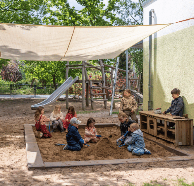 Eine Gruppe von Kindern spielt gemeinsam in einem großen Sandkasten auf einem sonnigen Spielplatz unter einem Sonnensegel.