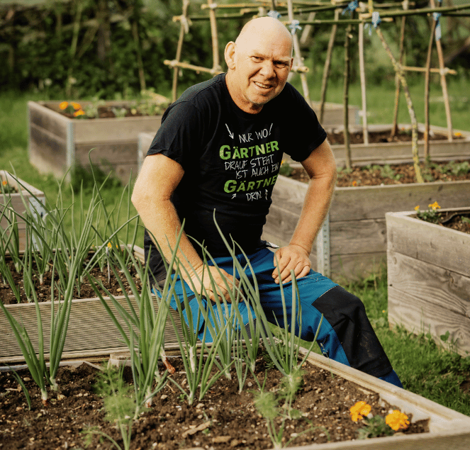 Jürgen Fritsche in einem Garten.Er sitzt auf einem Hochbeet mit Zwiebeln