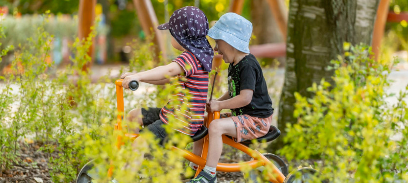 Zwei Kinder mit Sonnenhüten fahren im Schatten Dreirad