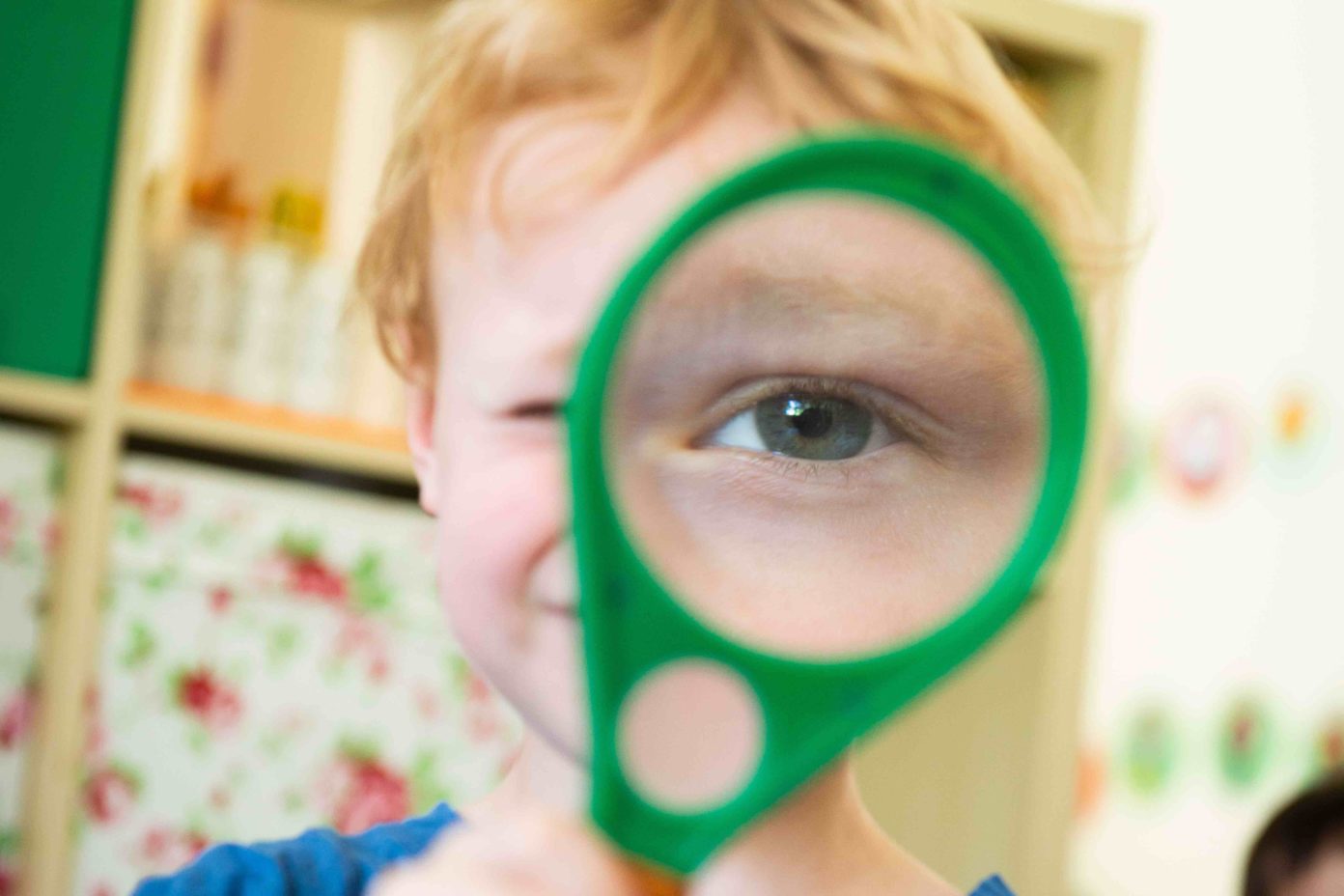 Portrait eines Kindes, das eine Lupe vors Auge hält, wodurch man das Auge im Vordergrund sehr groß sieht.