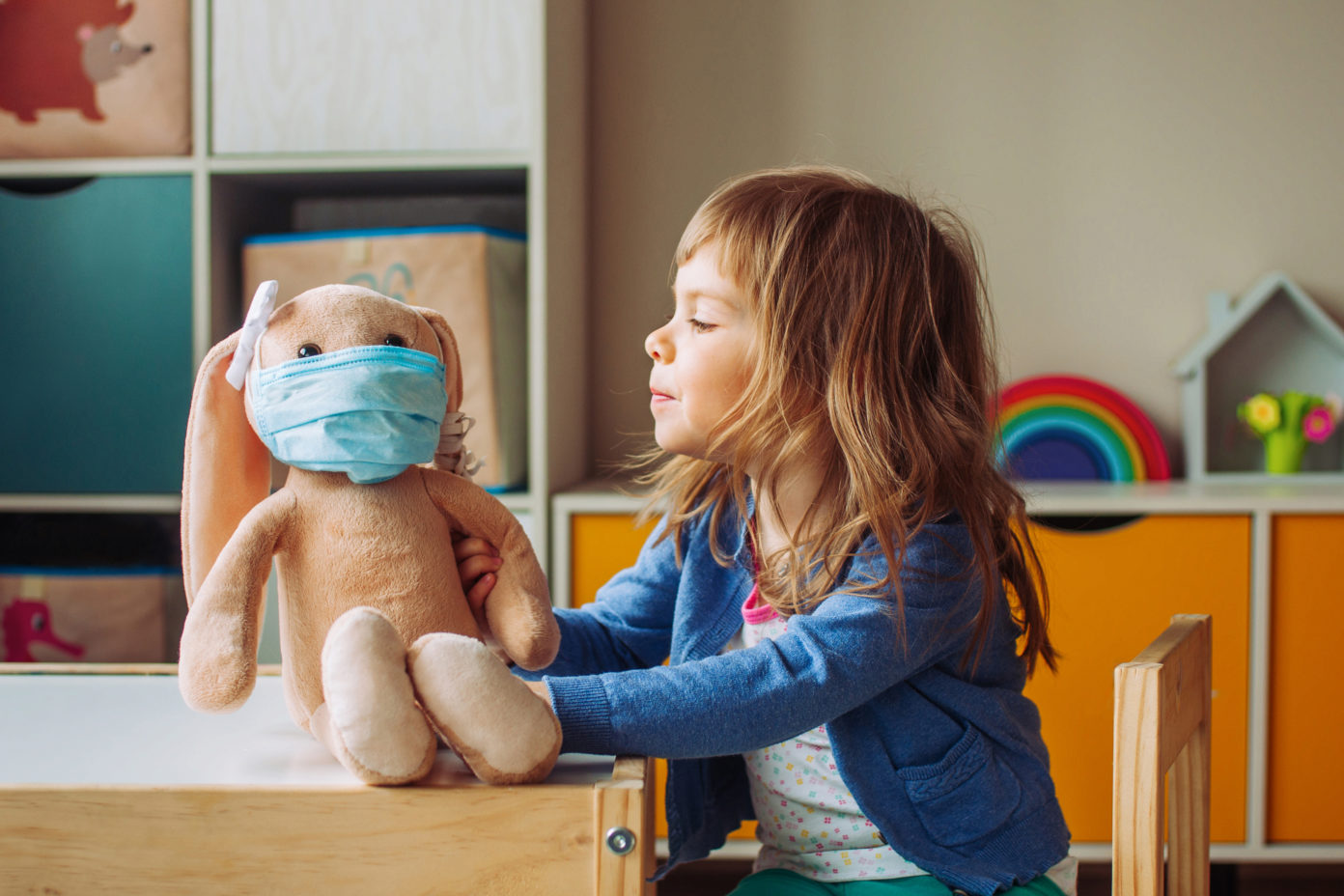 Ein kleines Mädchen spielt mit einem Stoffhasen, der eine medizinische Maske trägt. Sie sitzt an einem Tisch im Kinderraum.