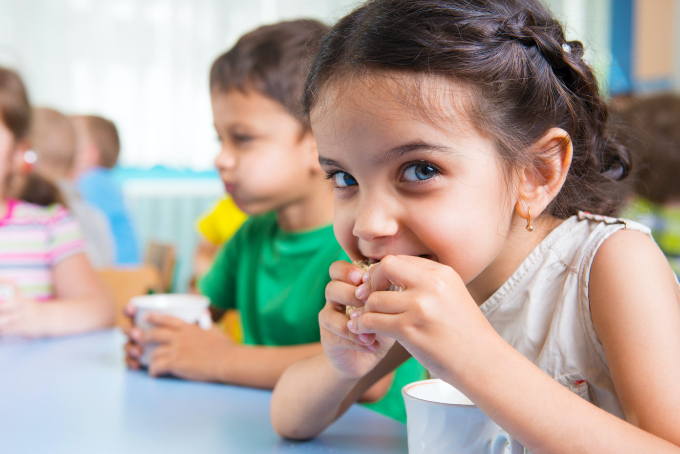 Ein kleines Mädchen sitzt an einem Kindergarten-Esstisch und isst einen Keks, der Junge neben ihr trinkt Milch.