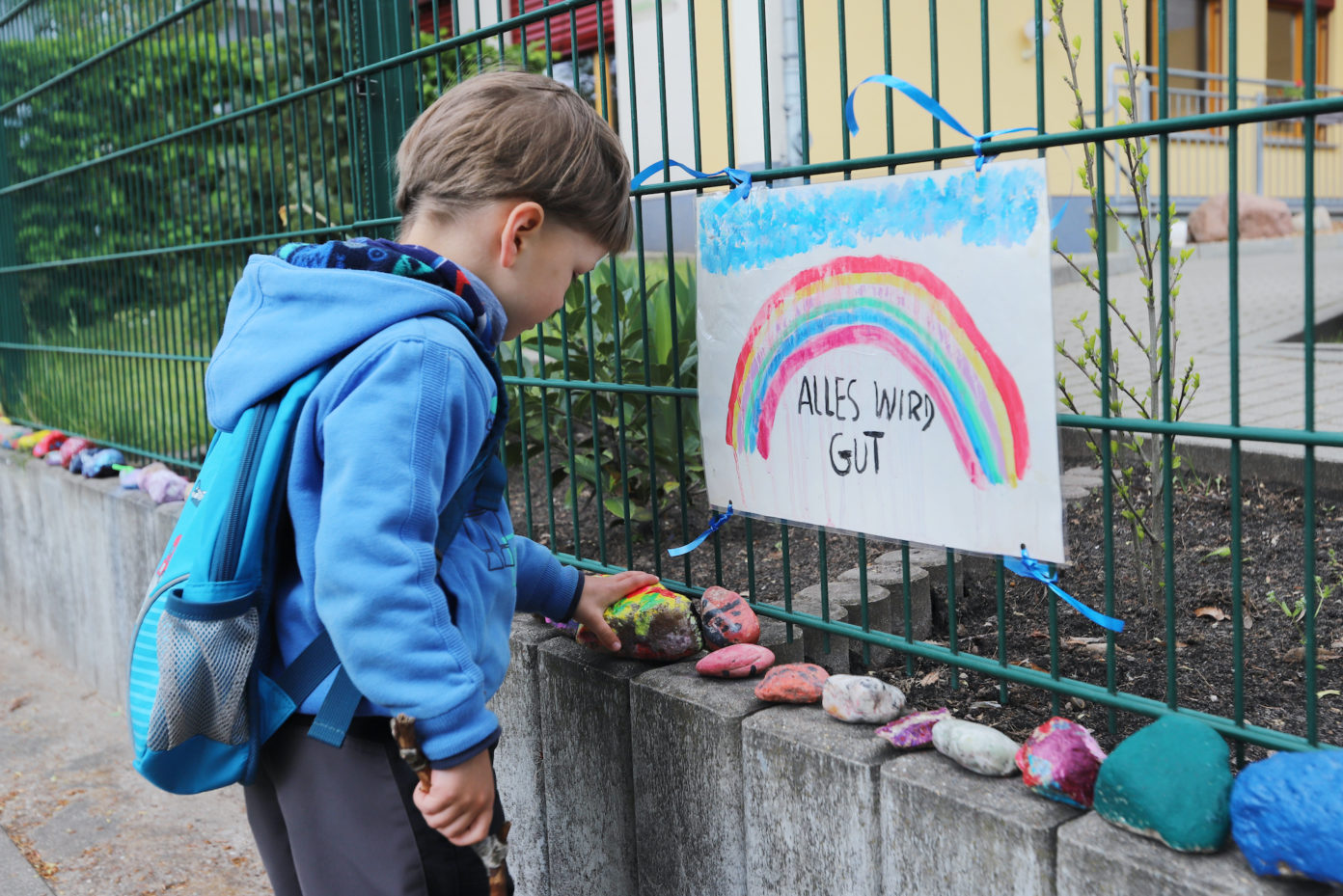 Ein kleiner Junge legt während der Corona-Zeit am Zaun vor seinem geschlossenen Kindergarten einen bemalten Stein unter ein Plakat mit Regenbogen, auf dem steht "Alles wird gut".