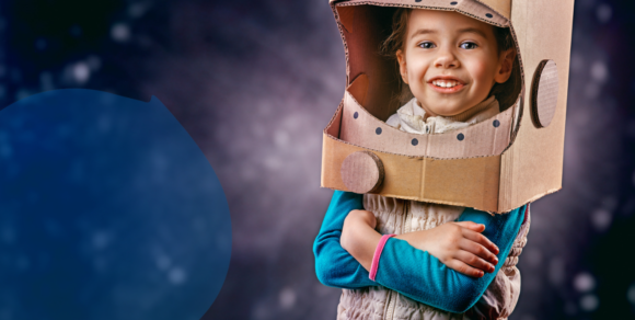 Foto eines etwa 5jährigen Mädchens, das einen Pappkarton wie einen Astronautenehlm über dem Kopf hat und vor einem Hintergrund steht, der das Weltall zeigt.