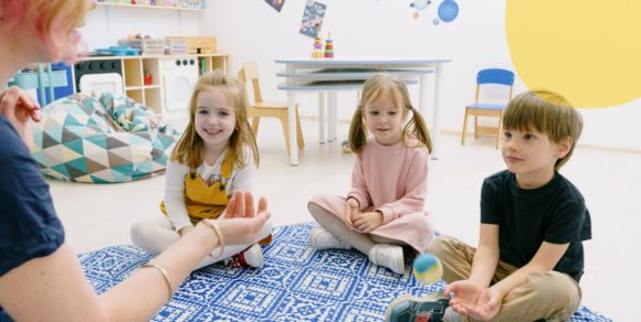 Kindergartenkinder sitzen im Halbkreis auf einem Spielteppich, vor ihnen eine Erzieherin