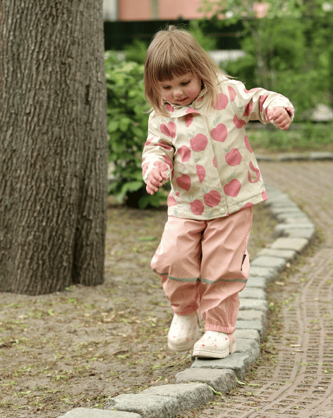 Ein kleines Mädchen mit Apfel-Jacke balanciert vorsichtig auf einer Steinmauer in einem Park im Freien.