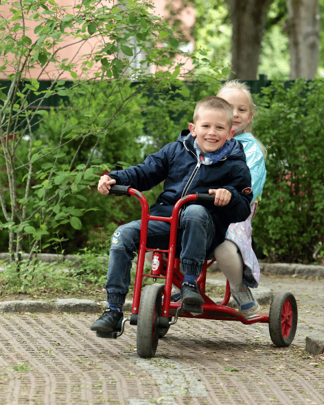 Ein Junge und ein Mädchen fahren gemeinsam auf einem roten Dreirad fröhlich durch einen grünen Garten im Freien.