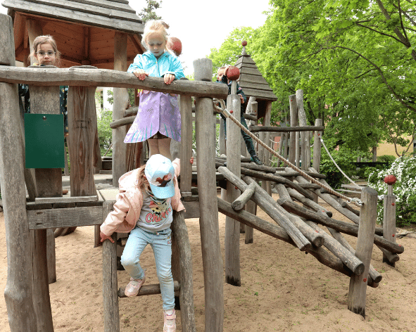 Mehrere Kinder spielen fröhlich auf einem hölzernen Klettergerüst auf einem sonnigen Spielplatz im Freien.