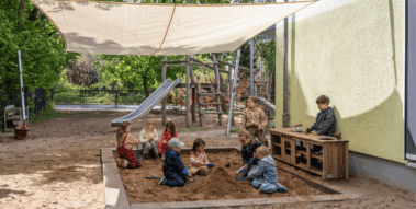 Eine Gruppe von Kindern spielt gemeinsam in einem Sandkasten auf einem Spielplatz unter einem großen Sonnensegel.