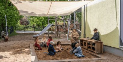 Eine Gruppe von Kindern spielt gemeinsam in einem Sandkasten auf einem Spielplatz unter einem großen Sonnensegel.
