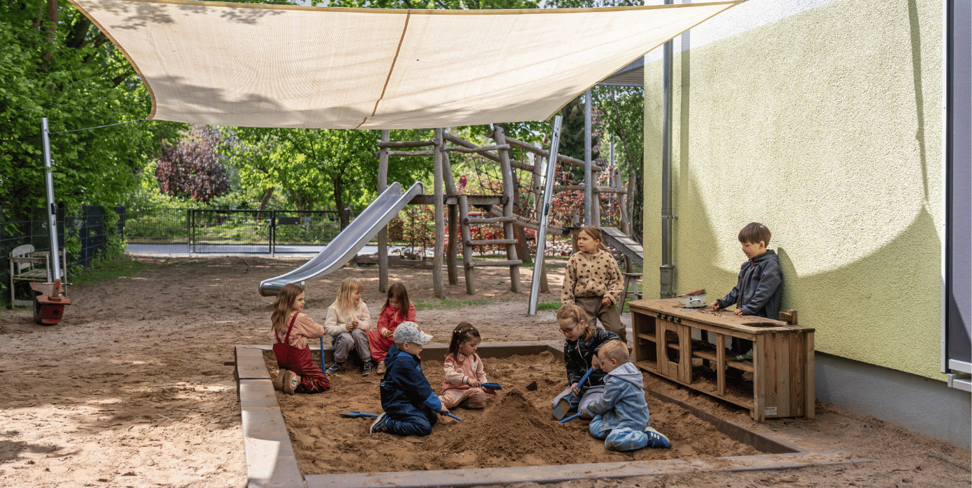 Eine Gruppe von Kindern spielt gemeinsam in einem Sandkasten auf einem Spielplatz unter einem großen Sonnensegel.