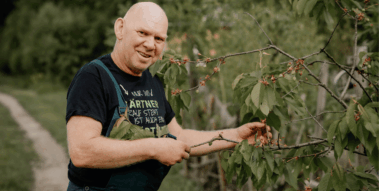 Ein freundlicher Gärtner, Jürgen Fritsche, mit Glatze und Latzhose steht in einem Garten und pflegt einen jungen Kirschbaum im Sommer.