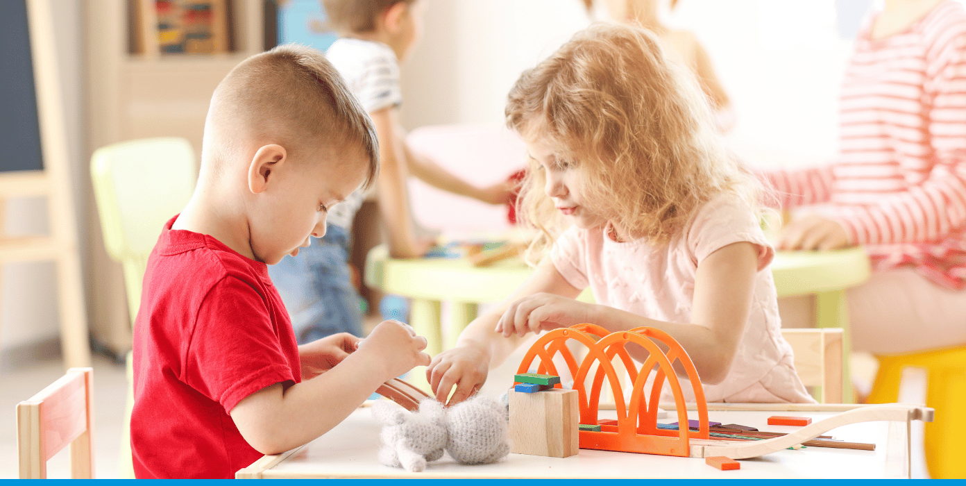 Ein Junge und ein Mädchen spielen konzentriert mit bunten Holzbausteinen und einer Brücke an einem Tisch im Kindergarten.